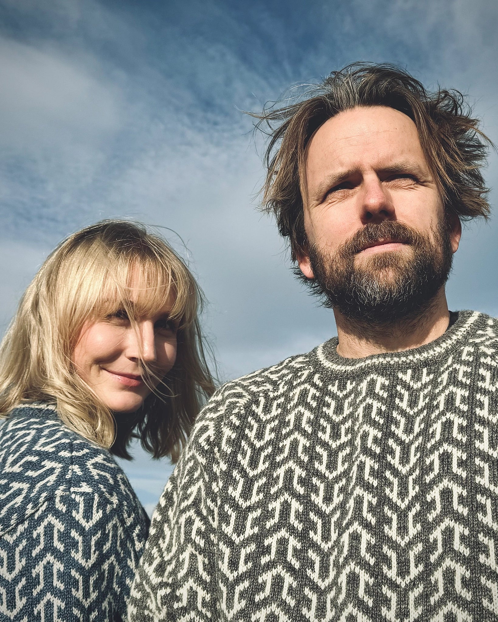 To personer i Nordvegr-gensere fra Vikingstrikk, en i grå og en i blå nyanser, ullgensere med nordisk mønster under åpen himmel.