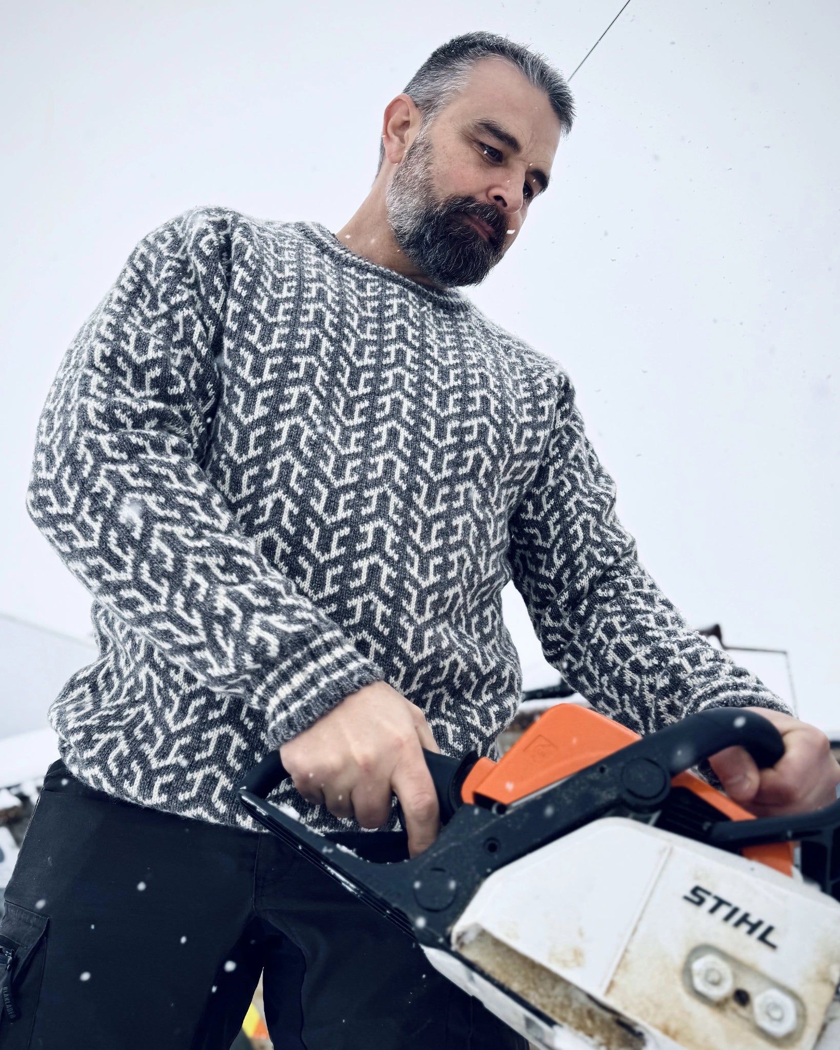 Mann i grå og hvit Nordvegr-genser fra Vikingstrikk, en allsidig ullgenser med nordisk mønster, brukt til arbeid ute i vinterkulde.