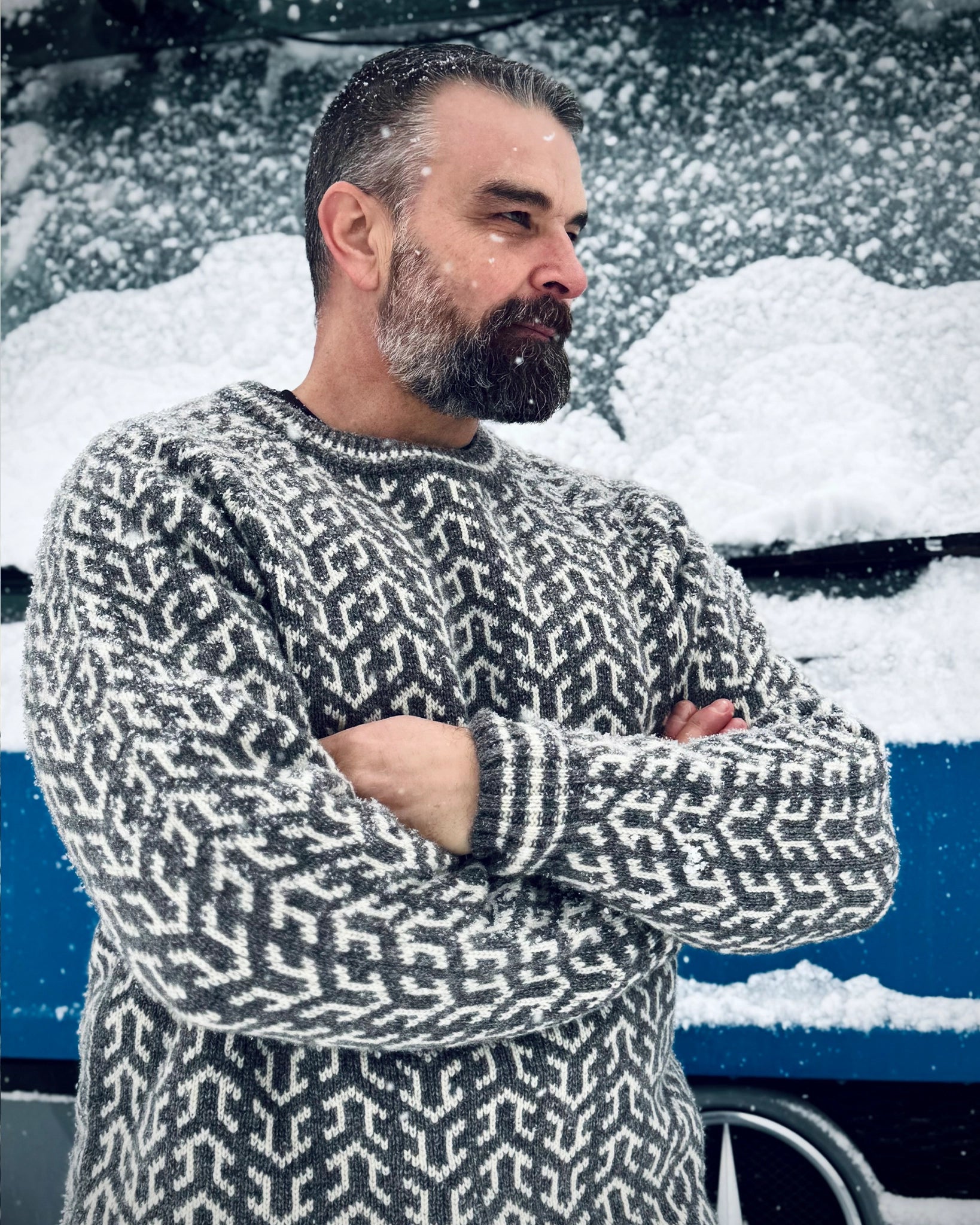Mann i grå og hvit Nordvegr-genser fra Vikingstrikk, en ullgenser med nordisk mønster, stående ute i snøvær med armene i kors.