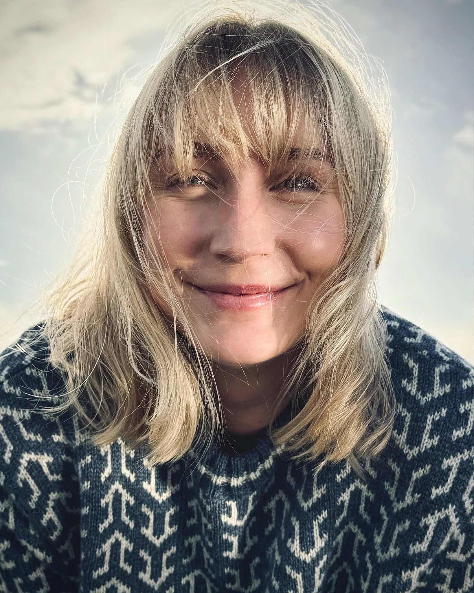 Nærbilde smilende kvinne i blå og hvit Nordvegr-genser fra Vikingstrikk, en ullgenser med nordisk mønster, fanget i naturlig lys.