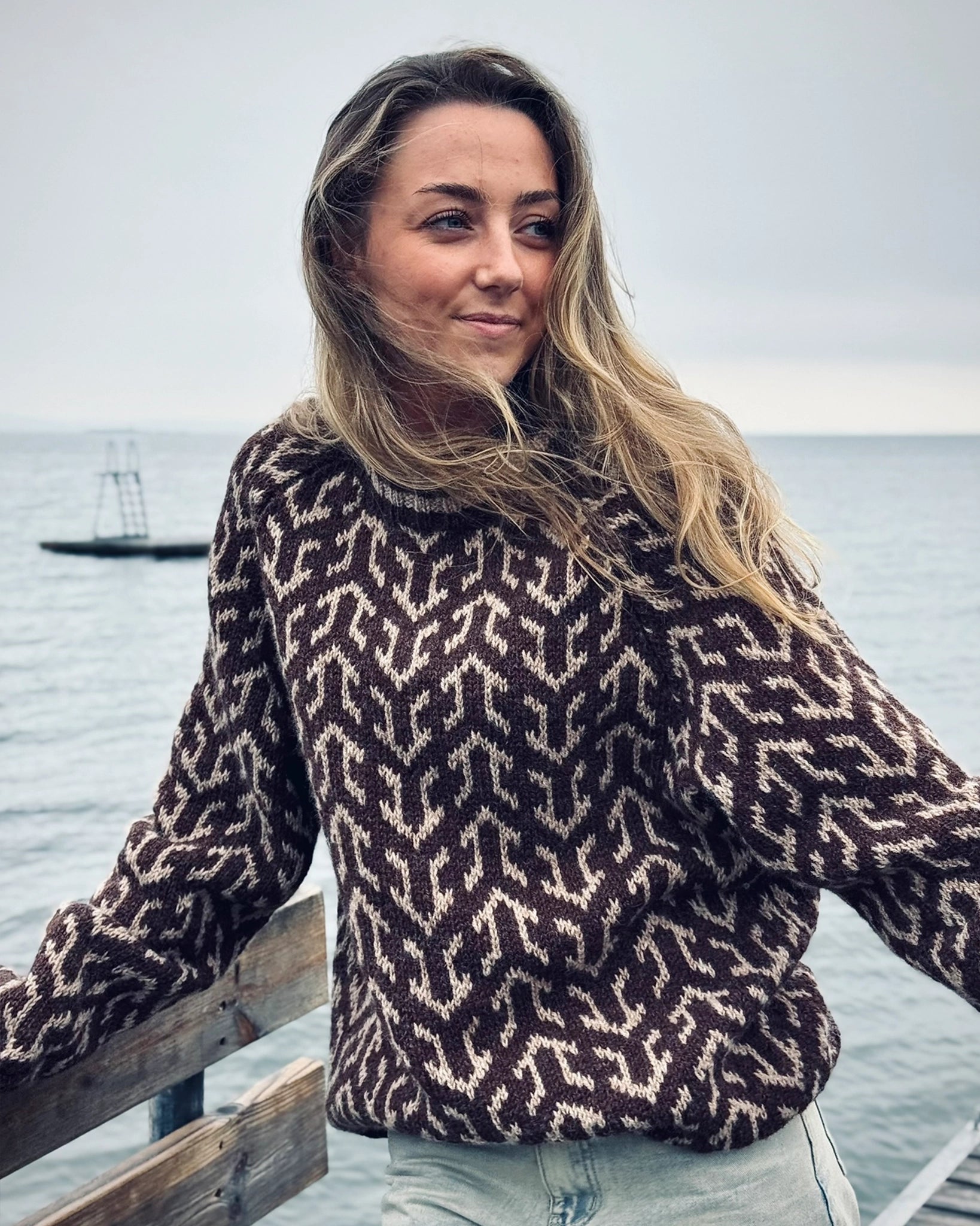 Kvinne ved sjøen iført Nordvegr-genser fra Vikingstrikk, en brun og hvit ullgenser med unikt mønster som selges som strikkepakke.