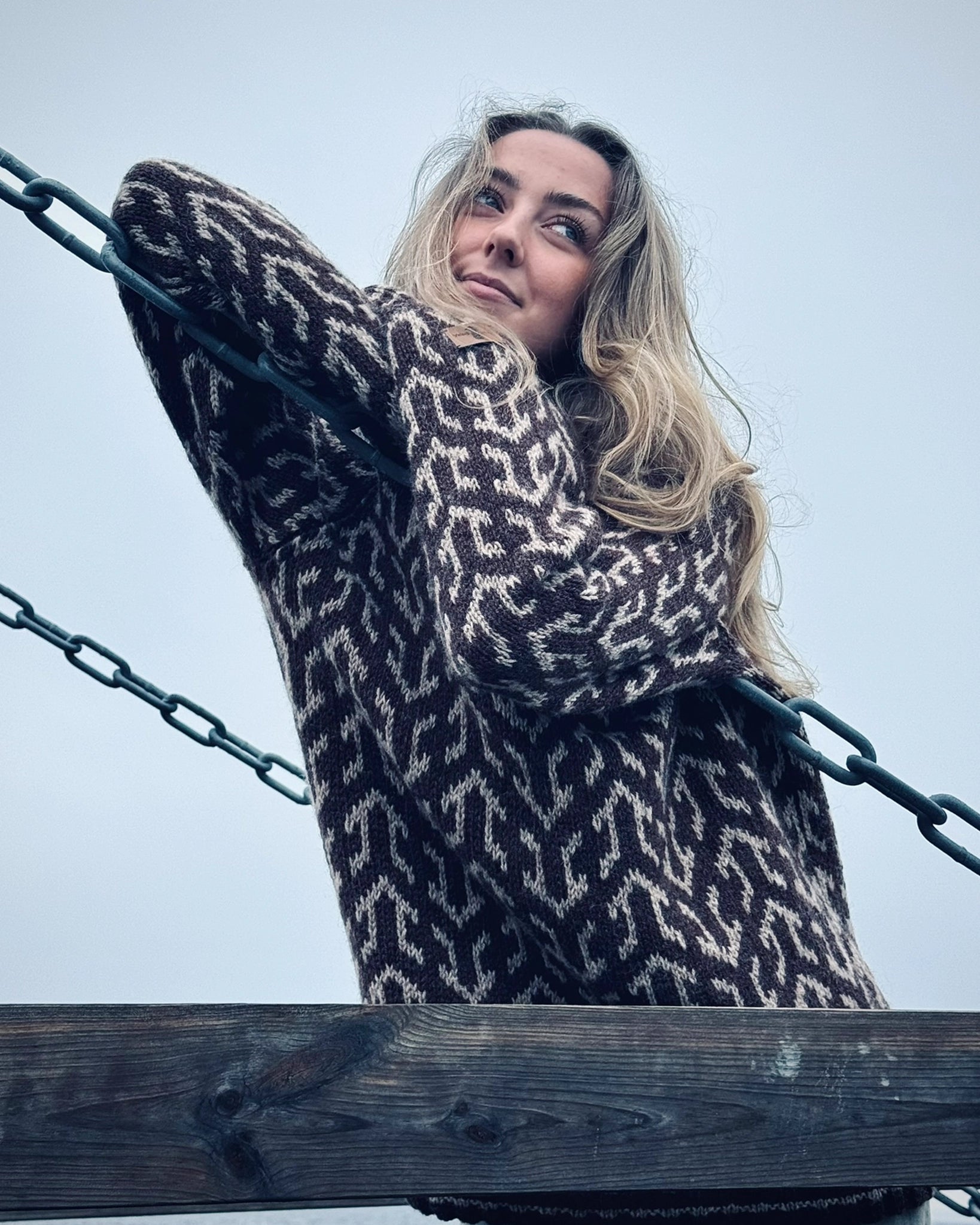 Kvinne i brun og hvit Nordvegr-genser fra Vikingstrikk, en ullgenser med unikt mønster som selges som strikkepakke, fotografert utendørs ved sjøen.