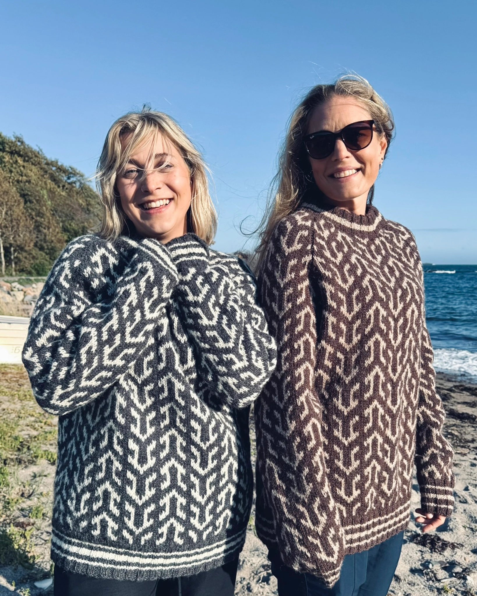 To kvinner smilende ved stranden iført Nordvegr-gensere fra Vikingstrikk, en i grå og hvit og en i brun og hvit, den unike genseren selges som strikkepakke.