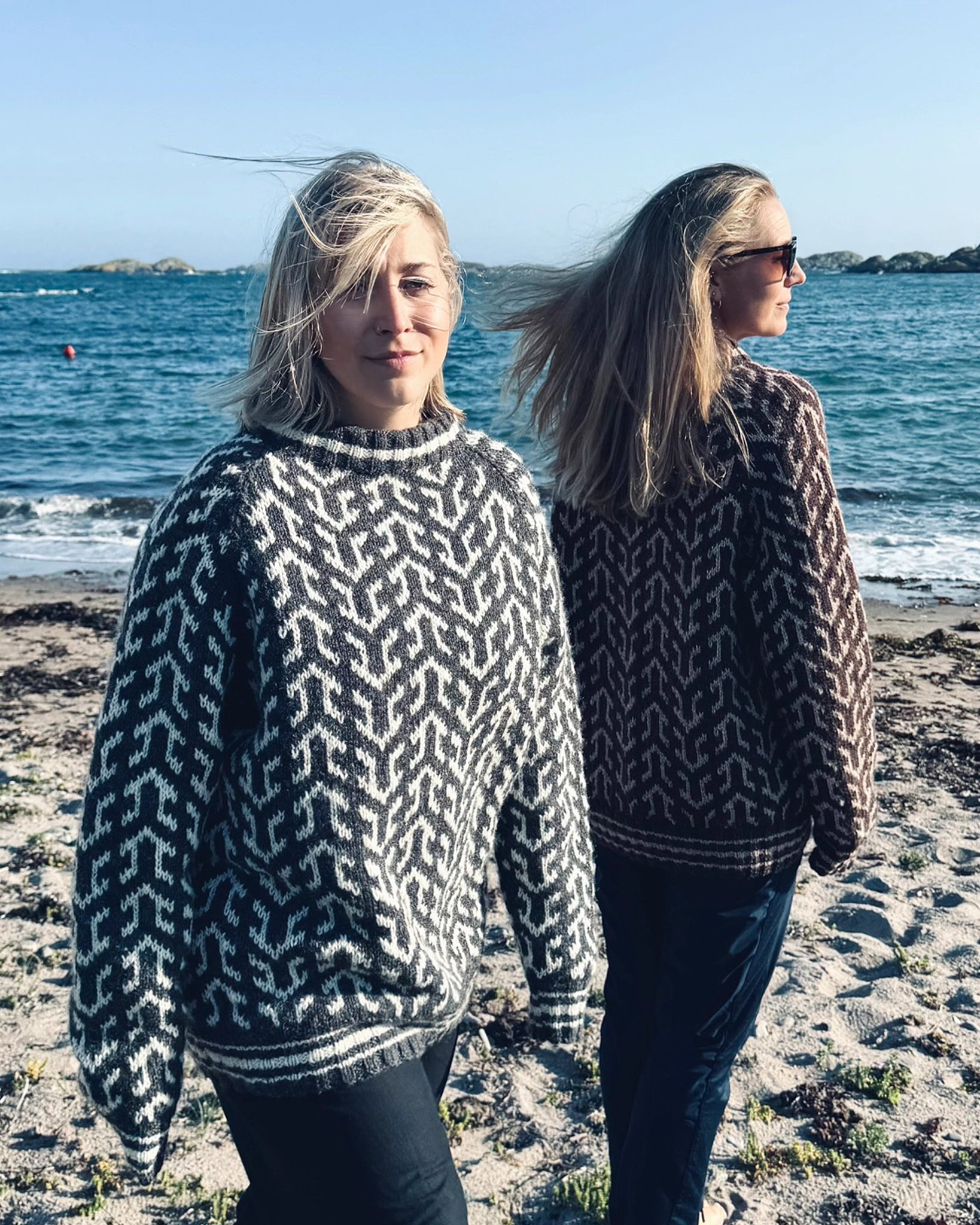 To kvinner ved stranden iført Nordvegr-gensere fra Vikingstrikk, en i grå og hvit og en i brun og hvit, ullgensere med unikt mønster. Selges som strikkepakke.