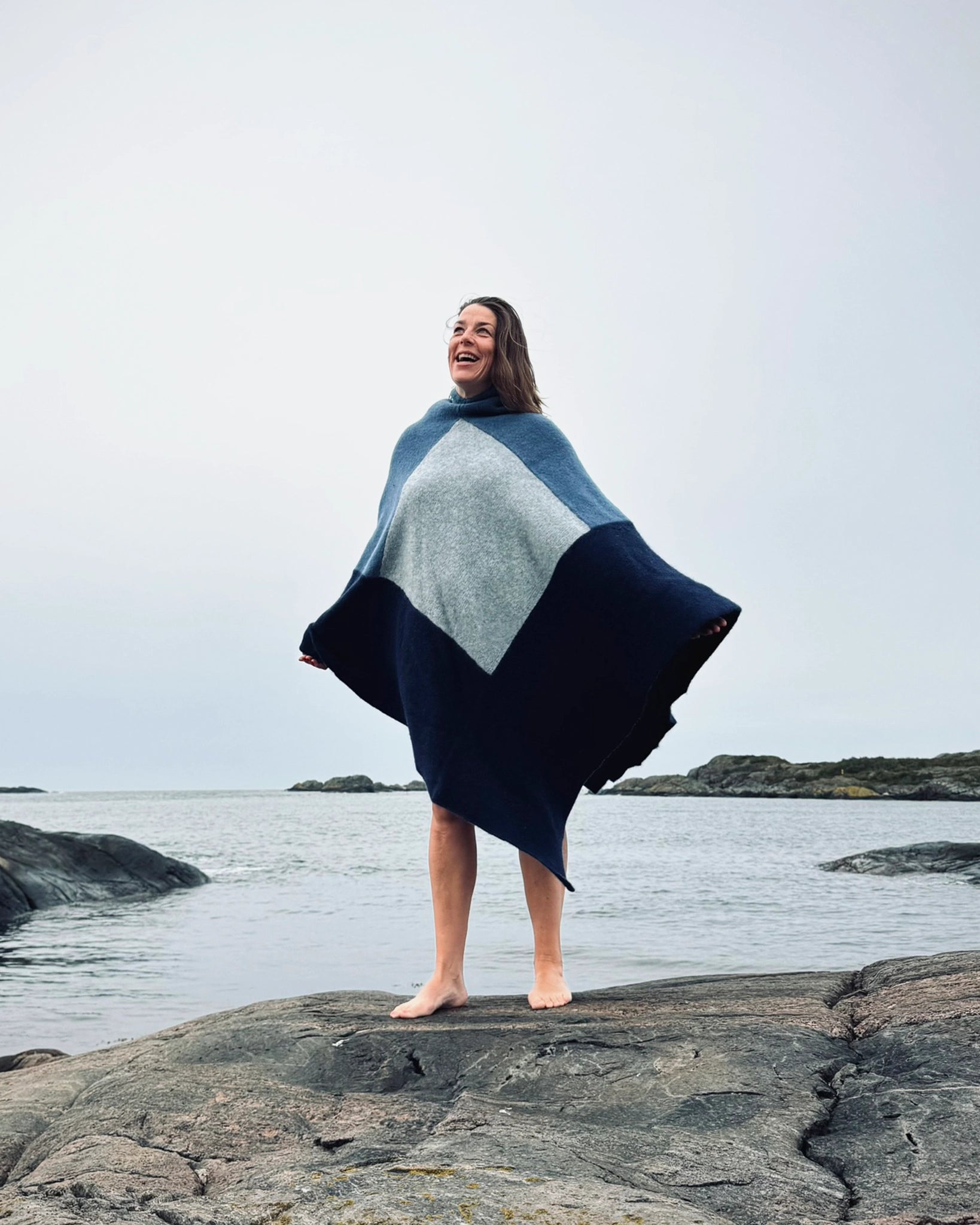 Totalt utsnitt av kvinne som står barføtt på svaberg ved sjøen iført blå og grå badeponcho fra Vikingstrikk, smilende med armene lett ut til siden.