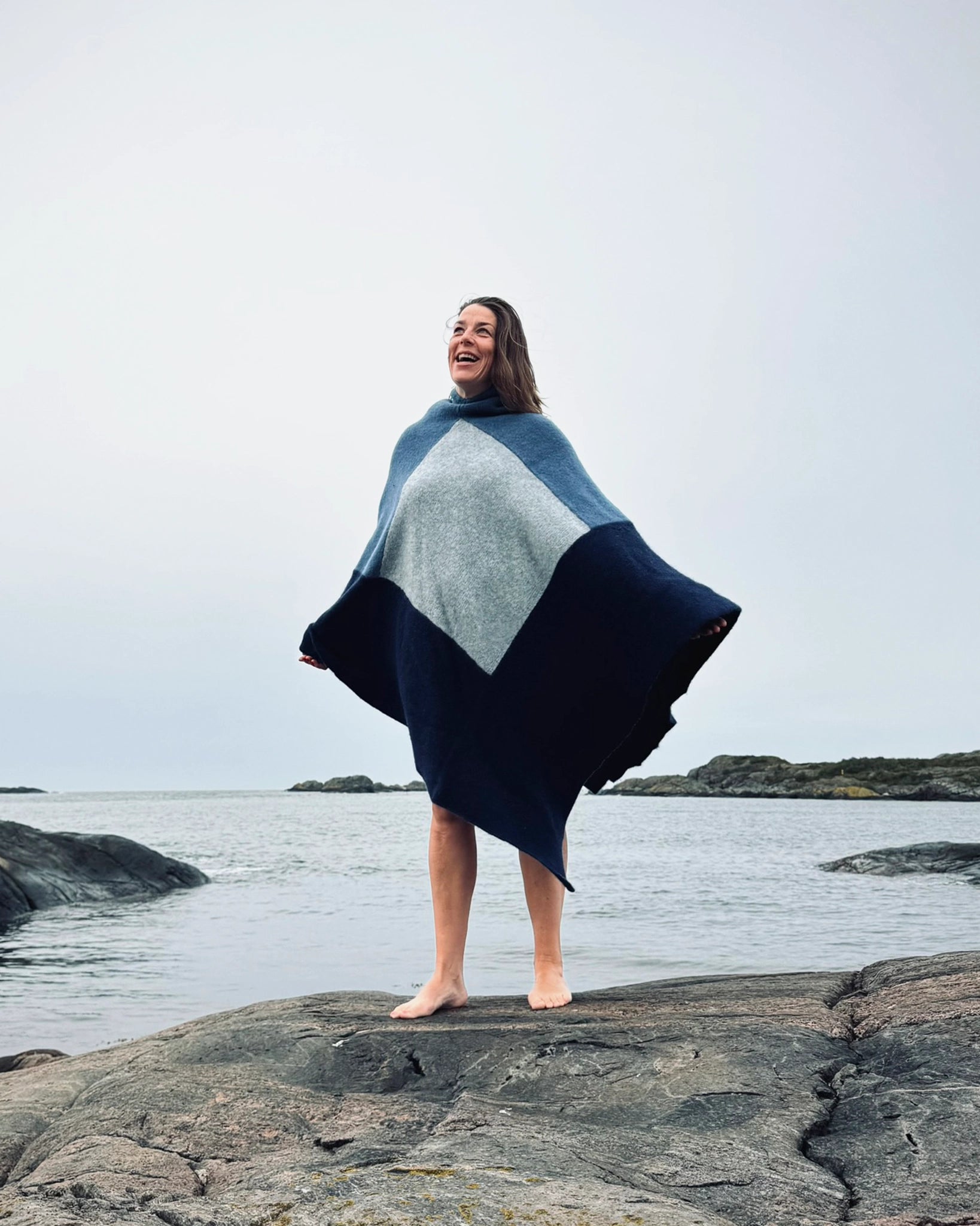 Totalt utsnitt av kvinne som står barføtt på svaberg ved sjøen iført blå og grå badeponcho fra Vikingstrikk, smilende med armene lett ut til siden.
