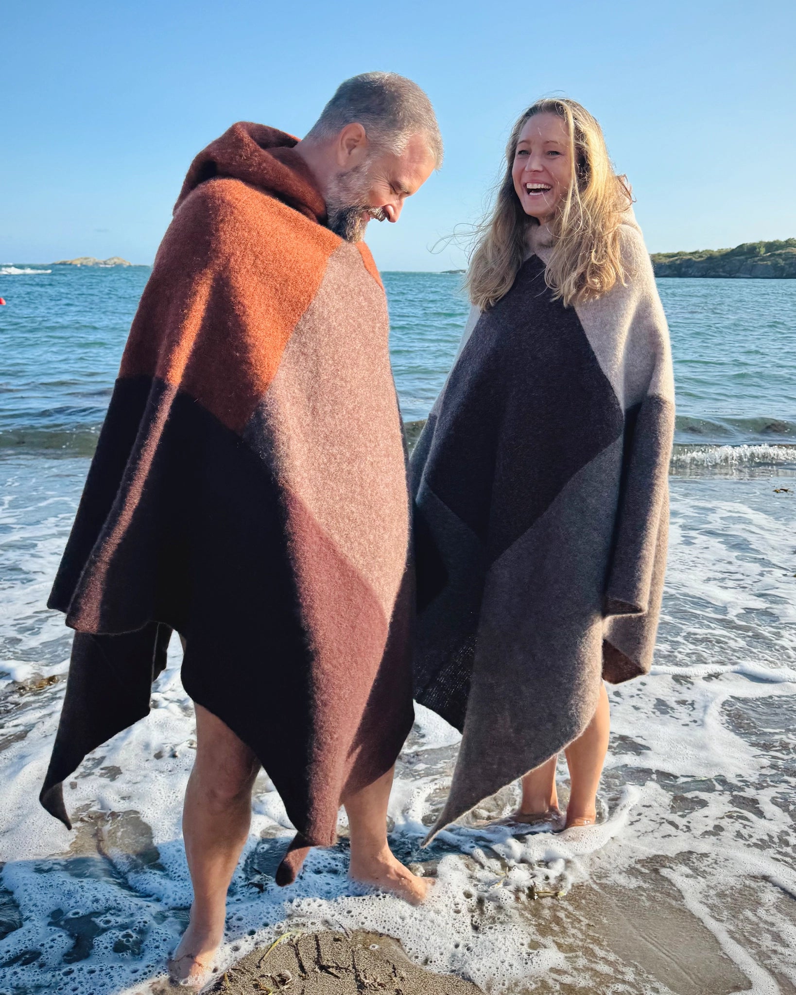 Totalt utsnitt av mann og kvinne som står barføtt i vannkanten iført ullponchoer fra Vikingstrikk i jordnære bruntoner, smilende og lekne ved stranden.