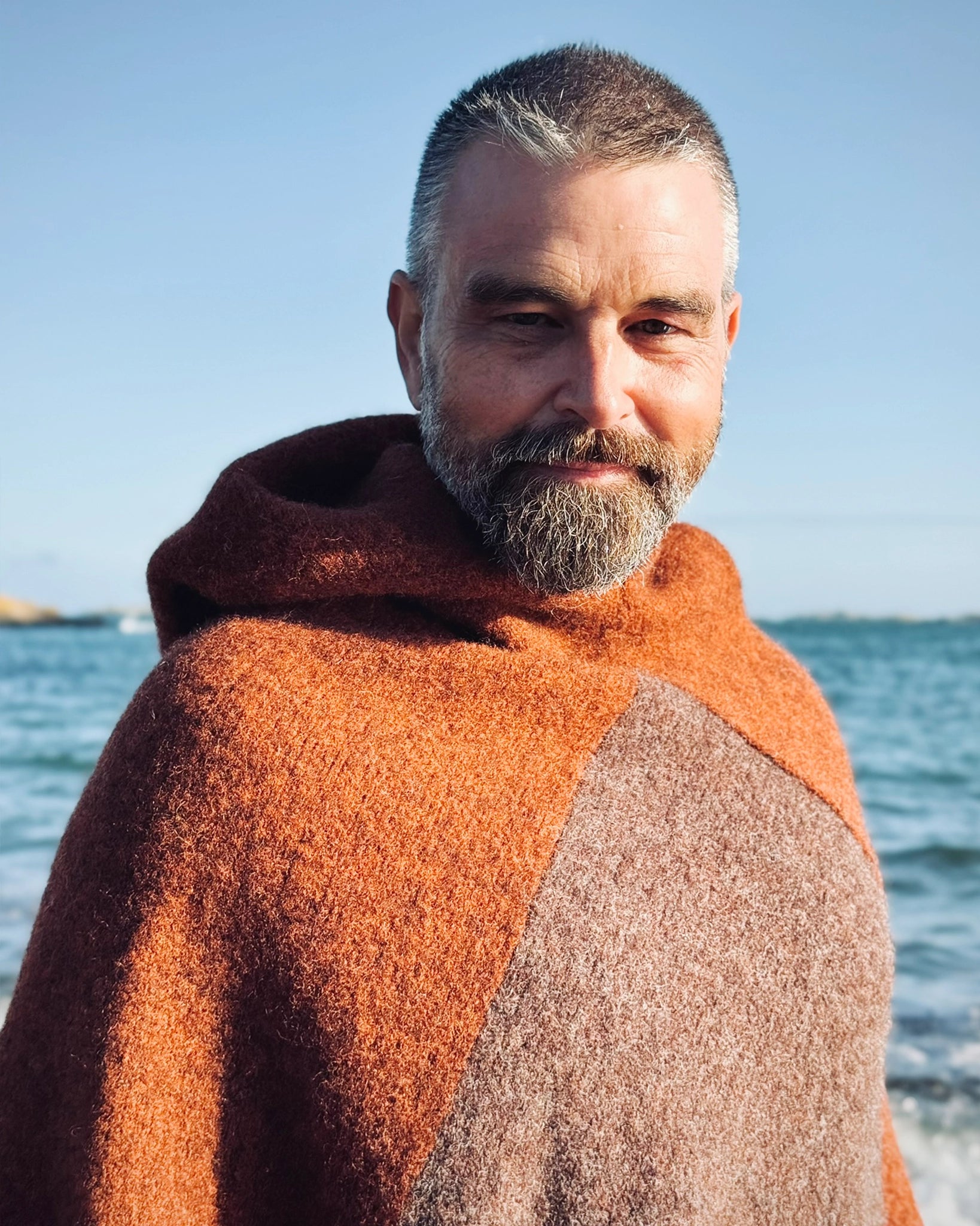 Halvnært portrett av mann ved sjøen iført ullponcho fra Vikingstrikk i rustbrune og beige nyanser, med hette og klassisk nordisk uttrykk.