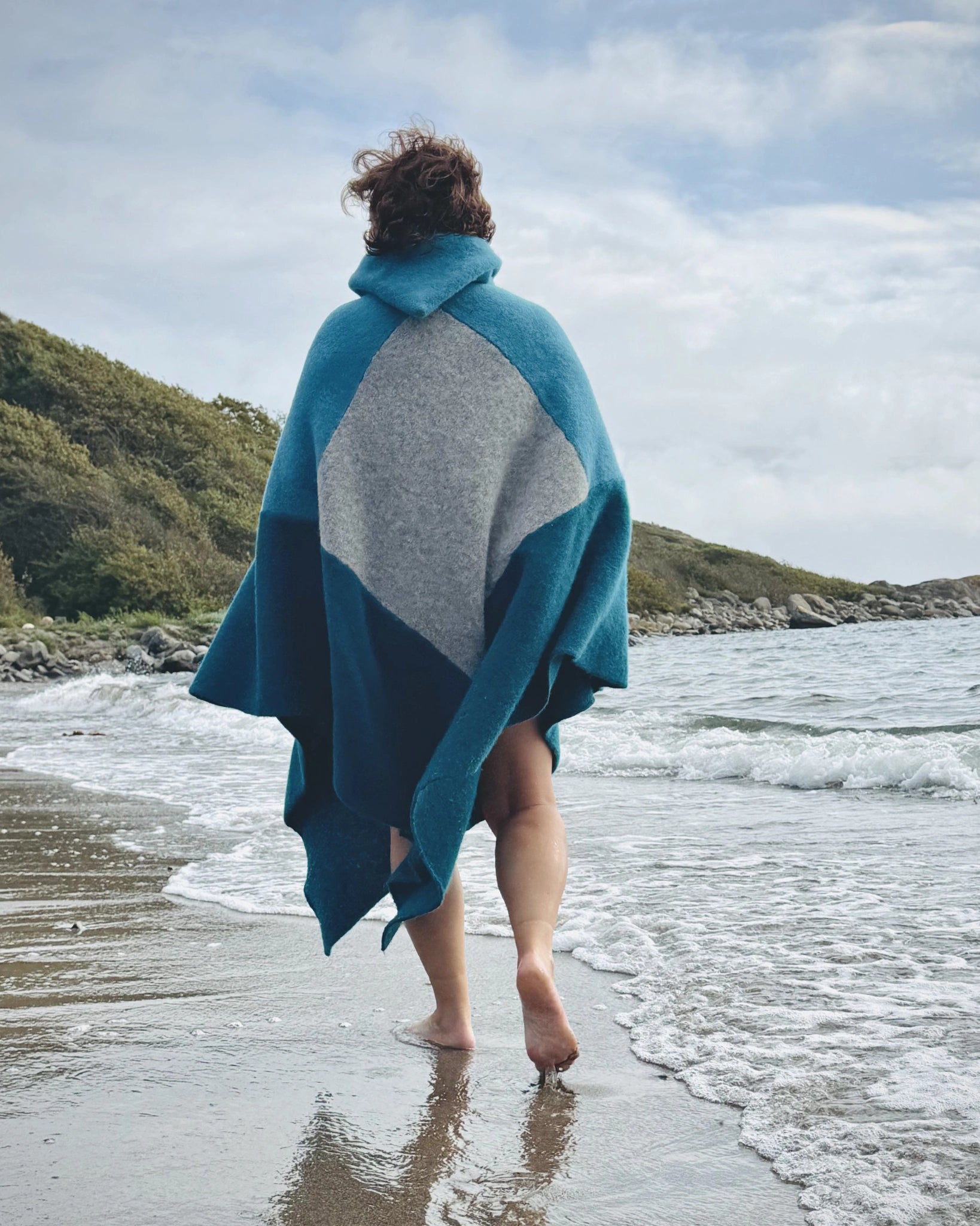 Totalt utsnitt bakfra av person som går barføtt i vannkanten iført blå og grå badeponcho fra Vikingstrikk, med natur og bølger i bakgrunnen.