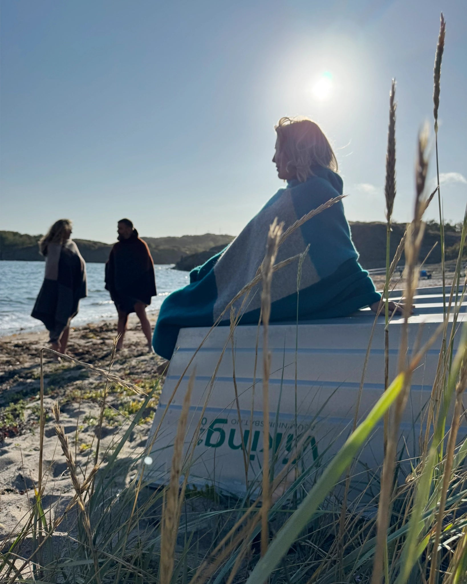 Strandscene med tre personer iført badeponchoer fra Vikingstrikk, en kvinne i blå poncho sitter på en båt i solnedgang mens to andre går barføtt langs vannet.