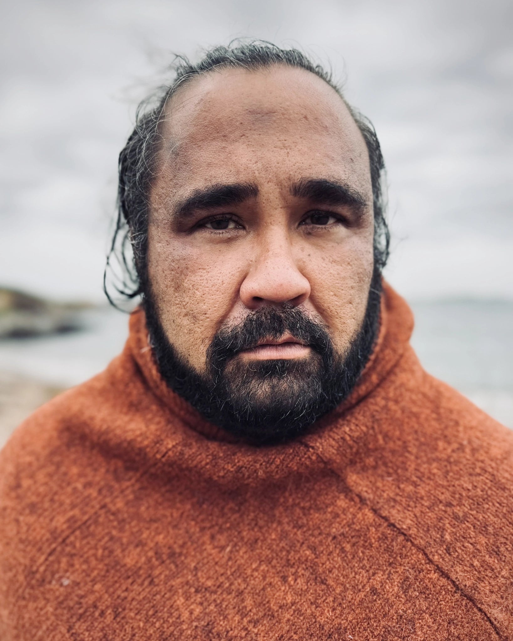 Halvnært portrett av mann ved sjøen iført rustrød Vikinghette fra Vikingstrikk, laget i ull med klassisk nordisk uttrykk.