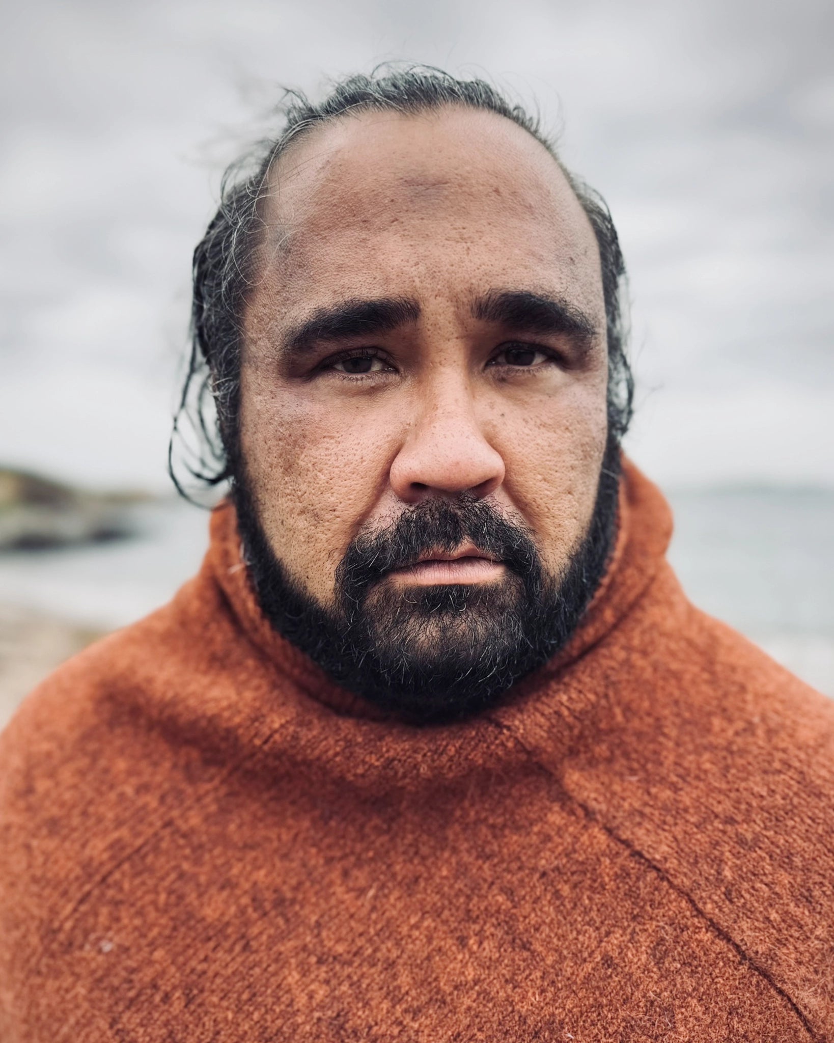 Halvnært portrett av mann ved sjøen iført rustrød Vikinghette fra Vikingstrikk, laget i ull med klassisk nordisk uttrykk.