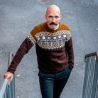 Nærbilde, mann i Kaizers Orchestra-genseren ser står i bunnen av en trapp og ser rett mot kamera. Ullgenseren er strikket i fargene brun, kamelbrun og natur. Nederste del av bolen er brun med mønster over brystet. Øverste del er kamelbrun og ermene er brune. Mønsteret er inspirert av logoen til Kaizers. Modellen er Helge Risa fra Kaizers Orchestra.