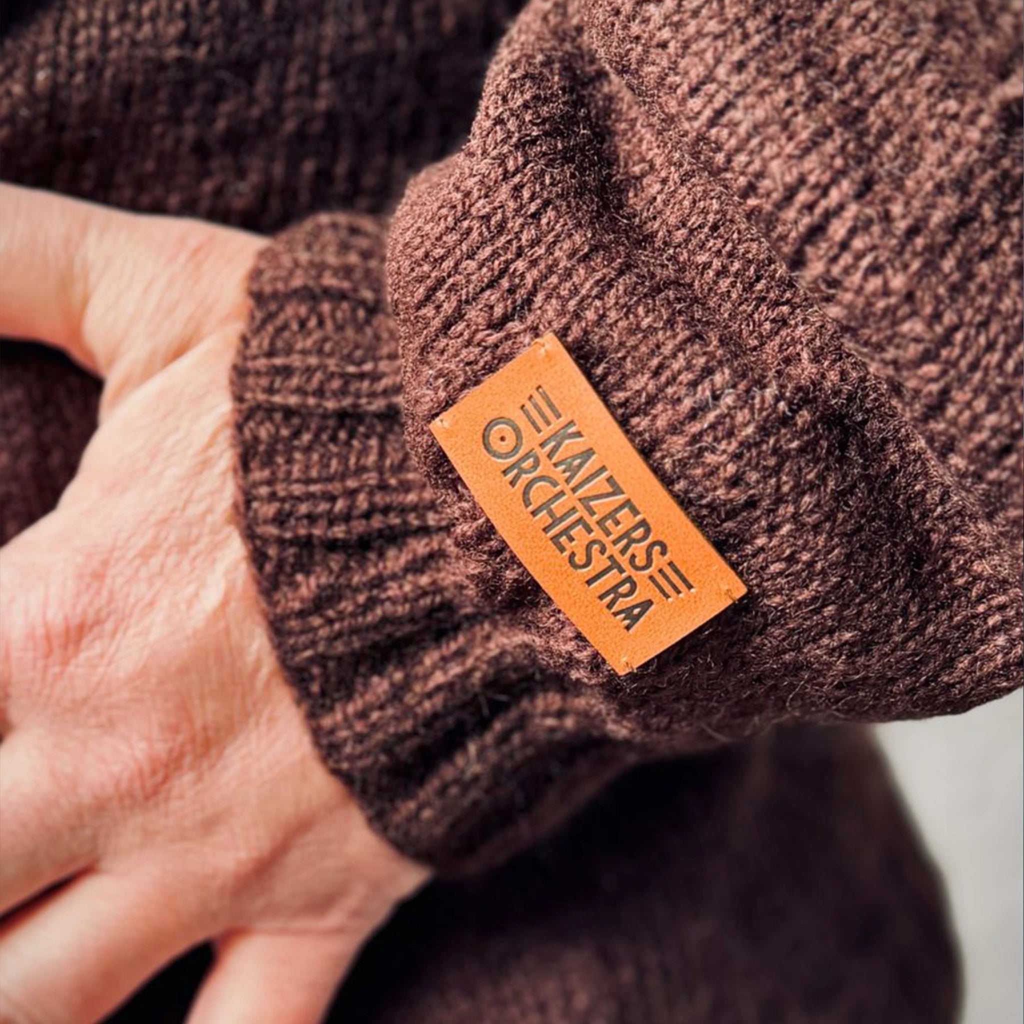 Ultranærbilde av nederste del av ermet på Kaizers Orchestra-genseren. Man ser hånden til modellen, vrangborden på genseren og nederste del av ermet. Fokuset i bildet er skinnlappen med logoen til Kaizers Orchestra.