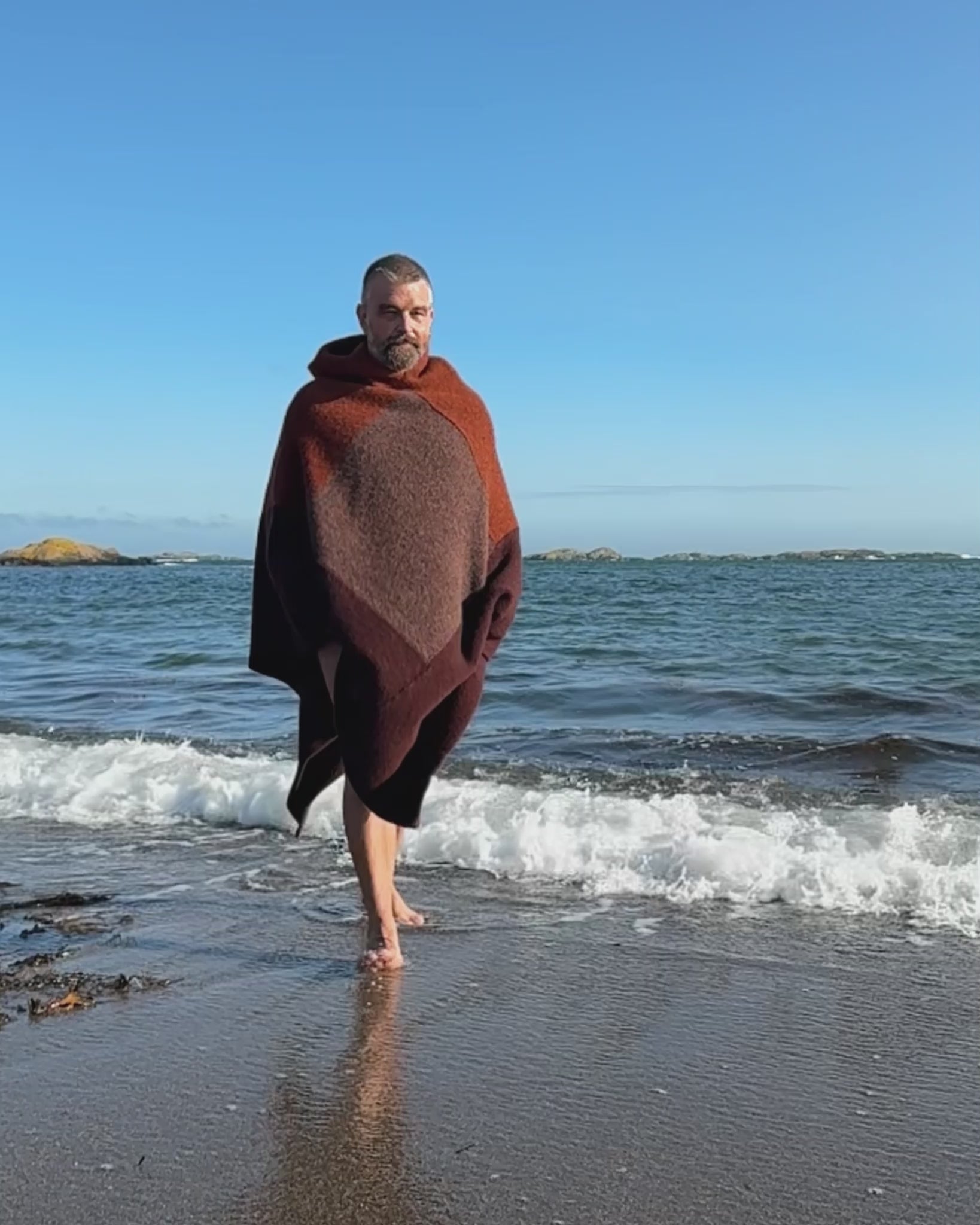 Totalt utsnitt av mann som går barføtt langs vannkanten iført rustrød og brun badeponcho fra Vikingstrikk, med sjøen og blå himmel i bakgrunnen.