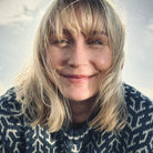 Nærbilde, kvinne i blå Nordvegr ullenser fra Vikingstrikk, ser rett i kameraet og smiler. Håret har blåst foran ansiktet hennes. 