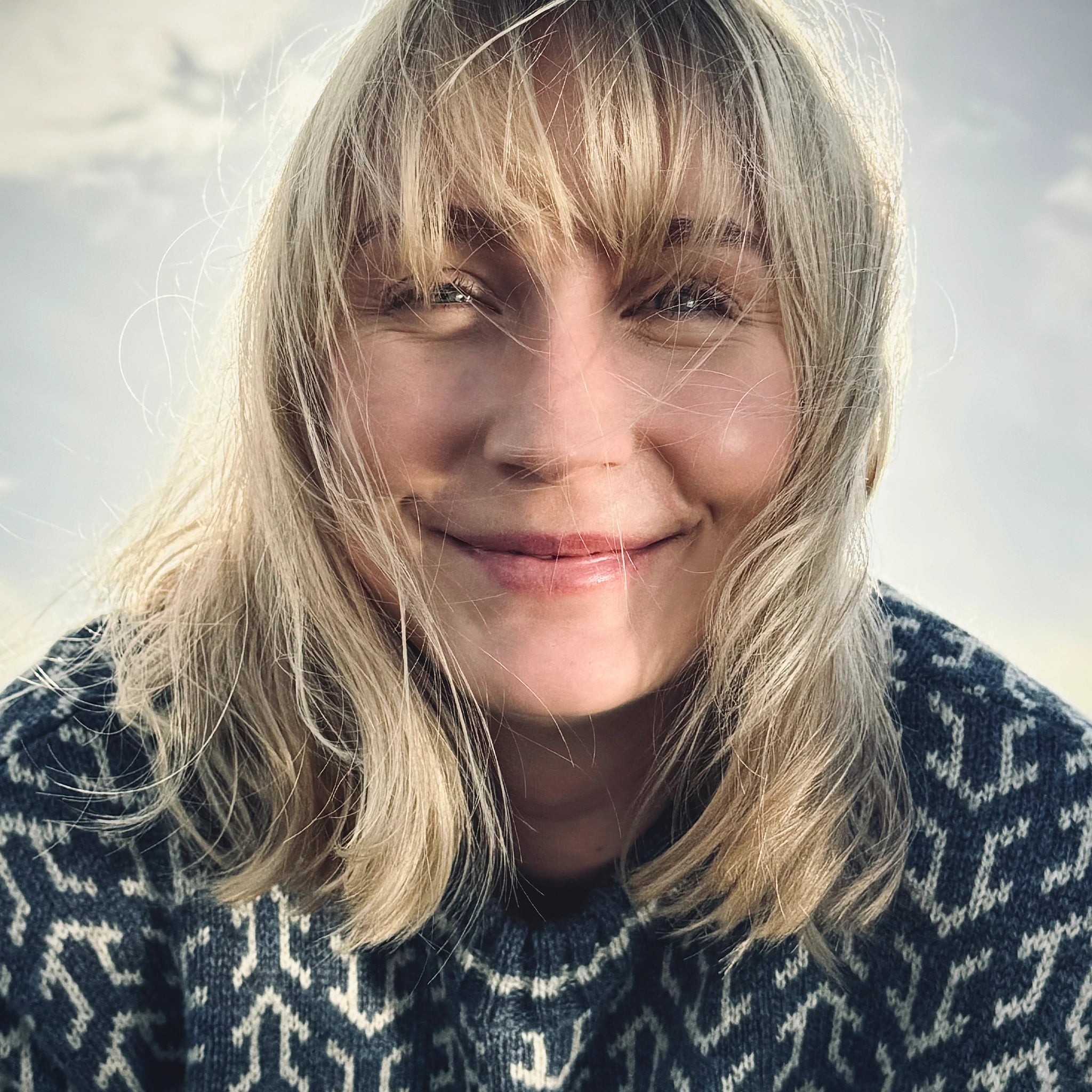 Nærbilde, kvinne i blå Nordvegr ullenser fra Vikingstrikk, ser rett i kameraet og smiler. Håret har blåst foran ansiktet hennes. 