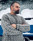Mann i grå og hvit Nordvegr-genser fra Vikingstrikk, en ullgenser med nordisk mønster, stående ute i snøvær med armene i kors.