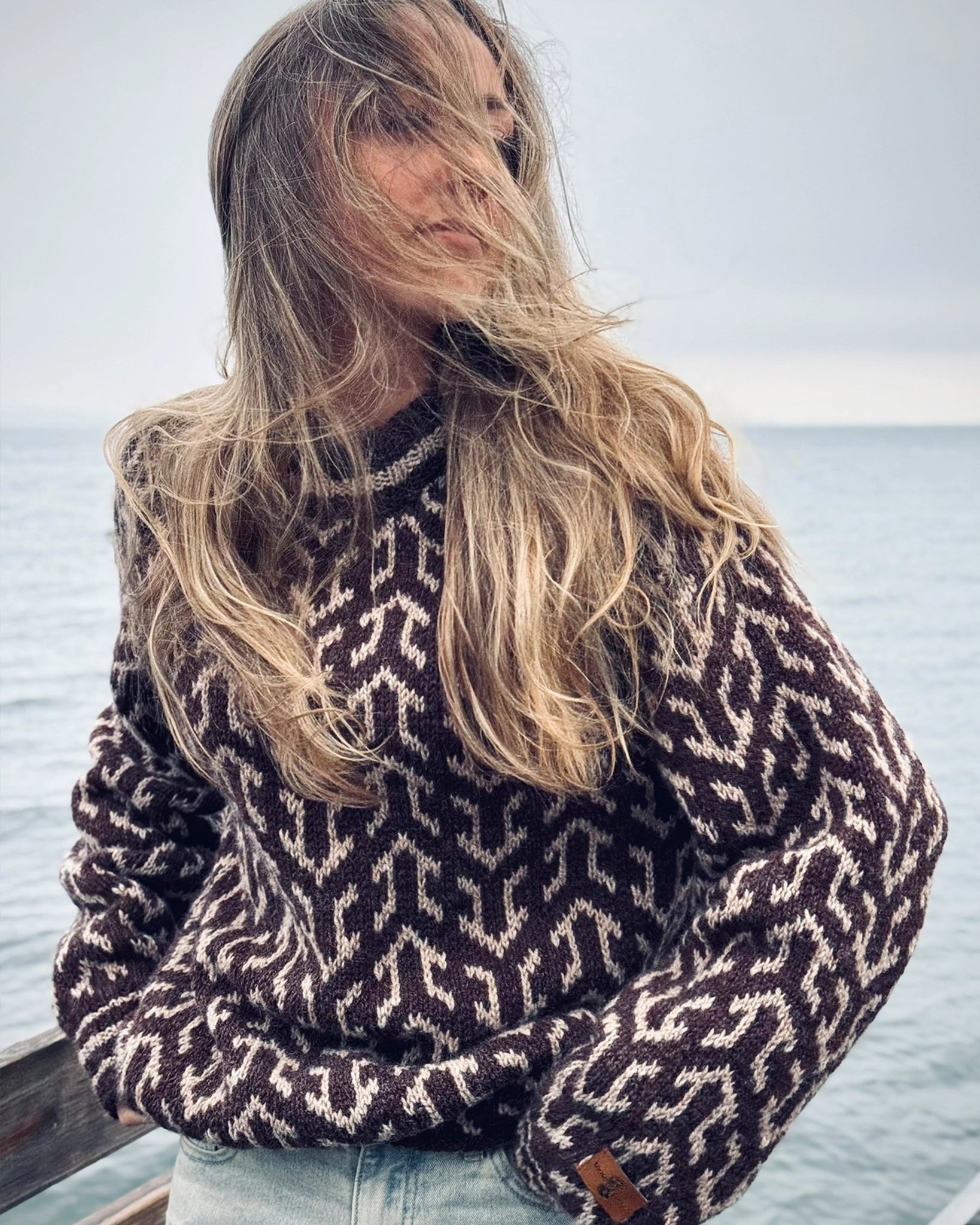 Halvtotal bilde av kvinne ved sjøen iført Nordvegr-genser fra Vikingstrikk, en brun og hvit mønstret ullgenser som selges som strikkepakke.