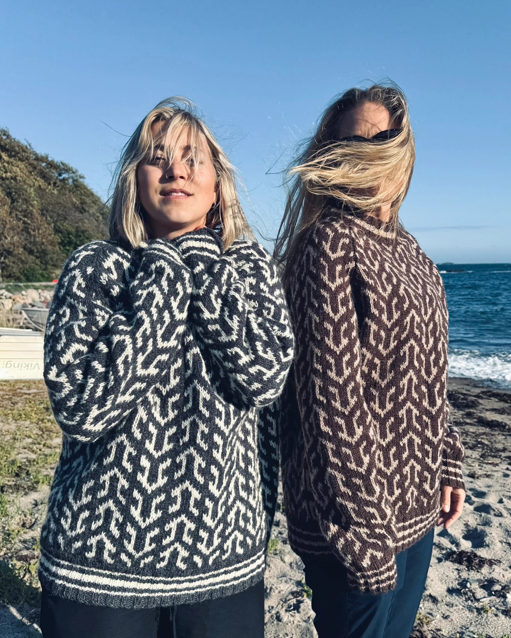 To kvinner ved stranden iført Nordvegr-gensere fra Vikingstrikk, en i grå og hvit og en i brun og hvit, den unike genseren selges som strikkepakke.