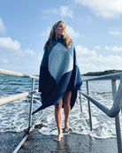 Totalt utsnitt av kvinne som går opp av vannet, iført ullponcho fra Vikingstrikk i blå nyanser, med blikket vendt ut av bilde.
