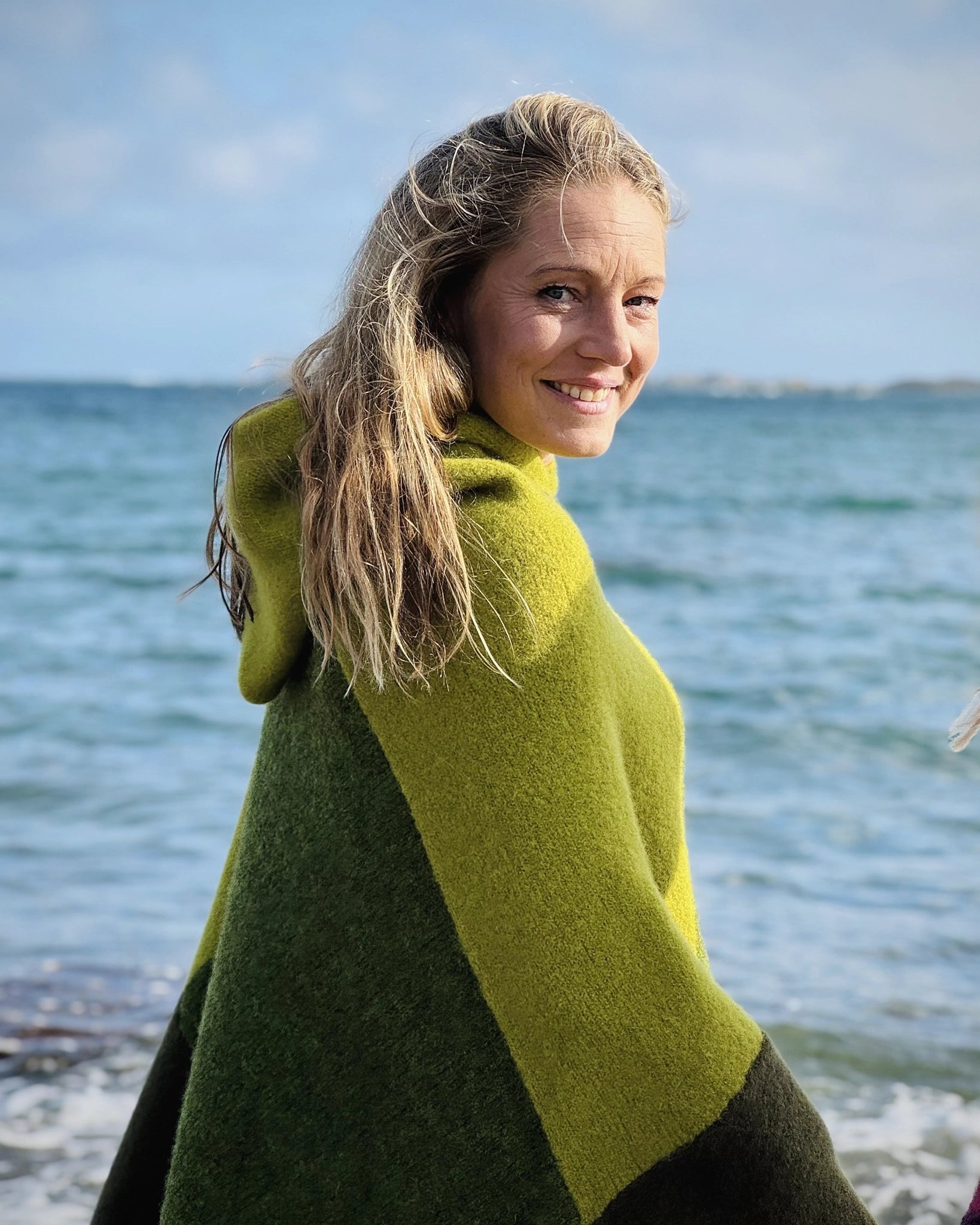 Halvnært portrett av kvinne ved sjøen iført ullponcho fra Vikingstrikk i grønne nyanser, smilende og vendt mot kamera i naturlig utendørslys.