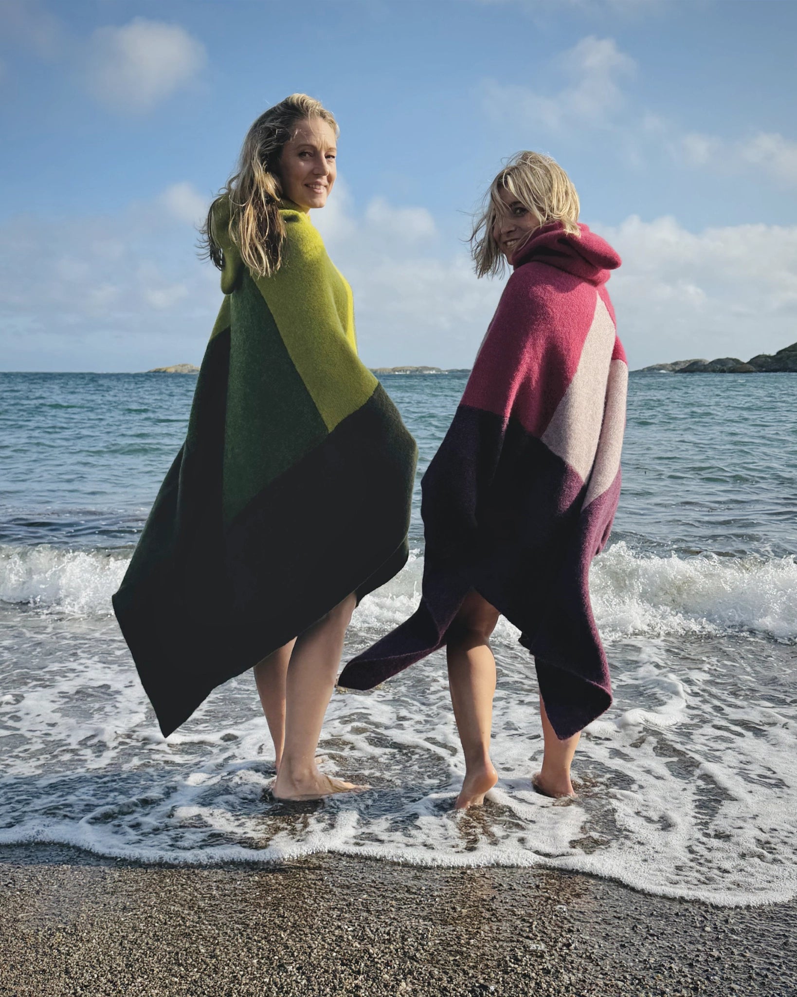 Totalt utsnitt av to kvinner som står barføtt i vannkanten iført ullponchoer fra Vikingstrikk, den ene i grønne nyanser og den andre i rosa og lilla, smilende ved stranden.