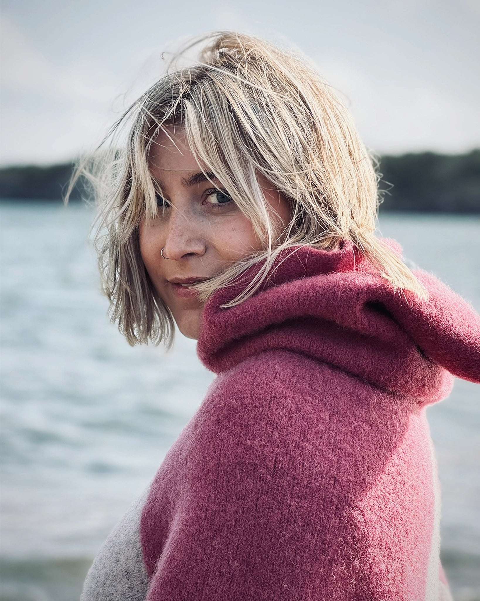 Kvinne i rosa ullponcho fra Vikingstrikk, halvnært bilde utendørs ved sjøen, med blikket rettet mot kamera.