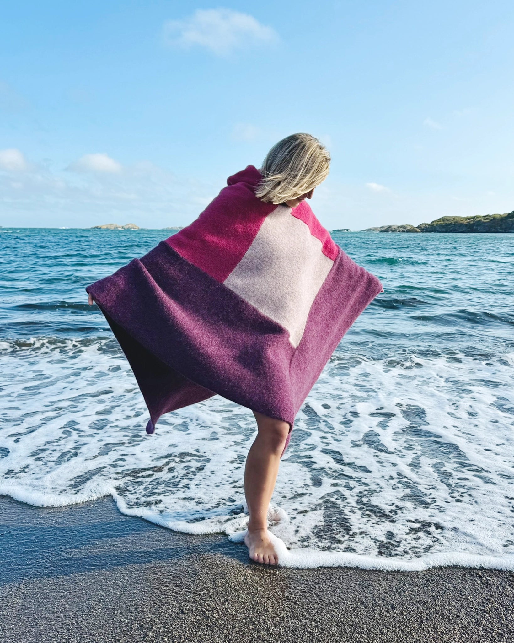 Totalt utsnitt av kvinne som står barføtt i vannkanten iført ullponcho fra Vikingstrikk i rosa og lilla nyanser, kvinnen løfter plagget ut med armene.