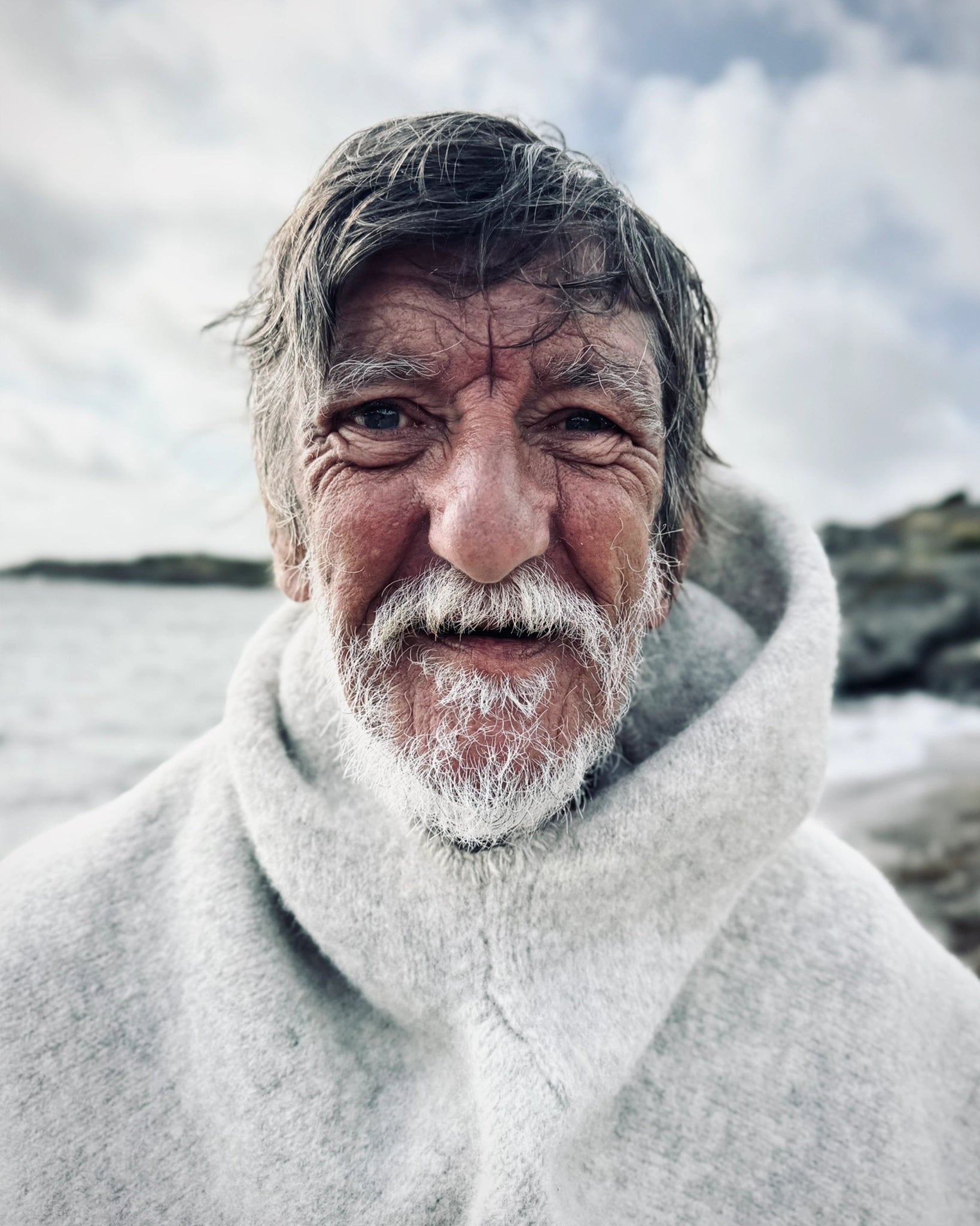 Nært portrett av eldre mann ved sjøen iført lys grå Vikinghette fra Vikingstrikk, med tydelige værbitt trekk og vennlig blikk. 